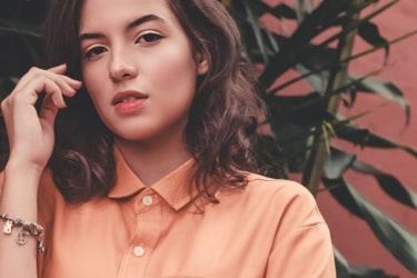 
          An elegant woman in a salmon-colored blouse stands gracefully against a lush backdrop of foliage.